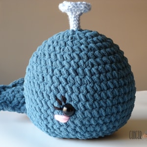 May include: A blue crocheted whale stuffed animal with a light blue blowhole and black eyes. The whale has a pink mouth and a white background.