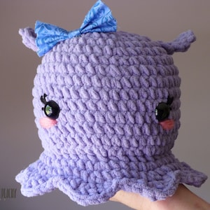 Puede incluir: Un juguete de peluche de pulpo de color morado tejido a crochet con un lazo azul, ojos negros y mejillas rosadas.