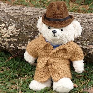 May include: A white teddy bear wearing a brown crocheted hat and a brown crocheted coat with a belt. The bear is sitting on a log in a forest setting.