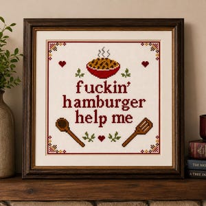 May include: A square cross-stitch embroidery in a dark wood frame featuring the text "fuckin' hamburger help me" in dark red letters. The design includes a red bowl of food, a wooden spoon, a spatula, and small red hearts on a white fabric.