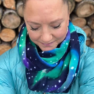 May include: A woman wearing a blue fleece jacket with a colorful infinity scarf featuring a galaxy print. The scarf is made of soft fleece and has a blue, green, purple, and white color scheme.
