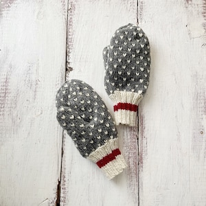 May include: A pair of grey knitted mittens featuring a white heart pattern. The cuffs are off-white with a red stripe. The mittens are displayed on a white, weathered wooden surface.