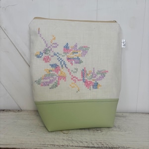 May include: A white linen zippered pouch with a green leather base. The pouch features a colourful embroidered floral design.