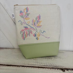 May include: A white linen zippered pouch with a green leather bottom. The pouch features a floral embroidery design in pink, yellow, blue, and green.