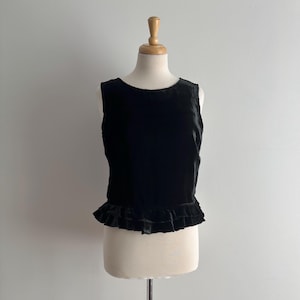 May include: A black velvet sleeveless top with a round neck and a frilled hem. The garment is displayed on a mannequin against a plain white backdrop. The fabric looks soft and drapes well, suggesting a comfortable fit.