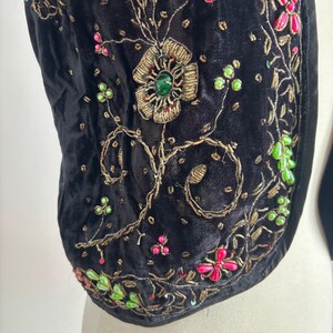 以下が含まれることがあります： 黒いベルベットのベストのクローズアップ。複雑な金色の刺繍とカラフルなビーズ細工が施されています。 デザインは、緑、ピンク、金色のアクセントが付いた花柄とつる植物のモチーフが特徴です。 ベストは曲線的な形状で、柔らかく質感のある外観です。