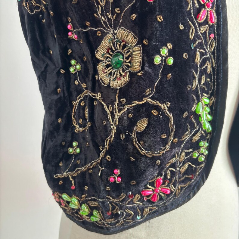 以下が含まれることがあります： 黒いベルベットのベストのクローズアップ。複雑な金色の刺繍とカラフルなビーズ細工が施されています。 デザインは、緑、ピンク、金色のアクセントが付いた花柄とつる植物のモチーフが特徴です。 ベストは曲線的な形状で、柔らかく質感のある外観です。
