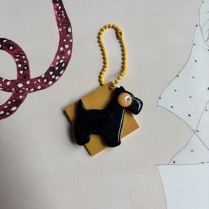 以下が含まれることがあります： 黄色いプラスチック製のタグが付いた黒いプラスチック製の犬のキーホルダー。犬は白い目と黄色い鼻をしています。