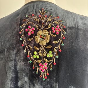 以下が含まれることがあります： 背面には刺繍の花柄が施された、濃いネイビーブルーのベルベットの衣服。刺繍は中央に金の花をあしらい、ピンクと緑のビーズのアクセントで囲まれています。デザインは金色の糸で縁取られています。