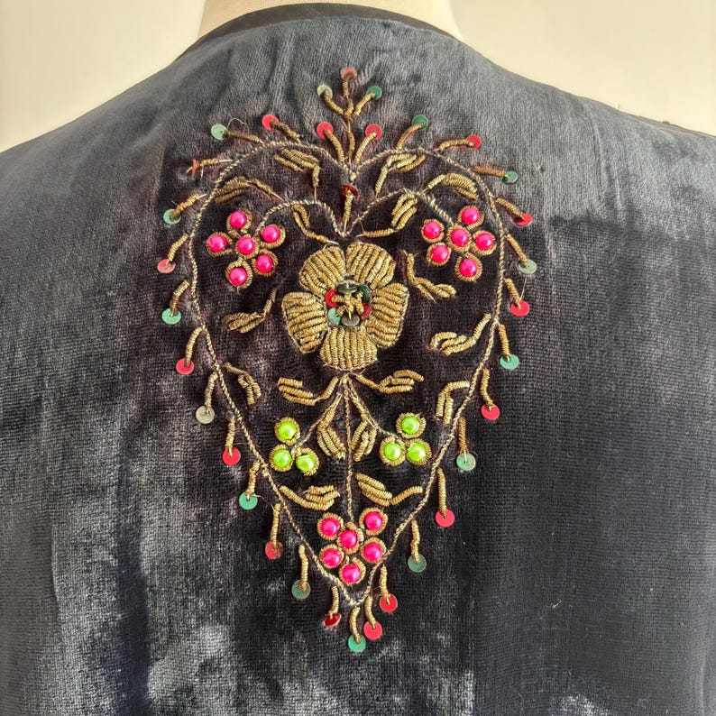 以下が含まれることがあります： 背面には刺繍の花柄が施された、濃いネイビーブルーのベルベットの衣服。刺繍は中央に金の花をあしらい、ピンクと緑のビーズのアクセントで囲まれています。デザインは金色の糸で縁取られています。