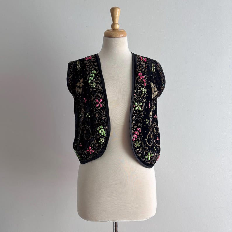 May include: A black velvet vest with pink, green, and gold floral embroidery. The sleeveless vest has an open front, shown on a mannequin. The embroidery forms a decorative border along the edges and around the floral patterns.