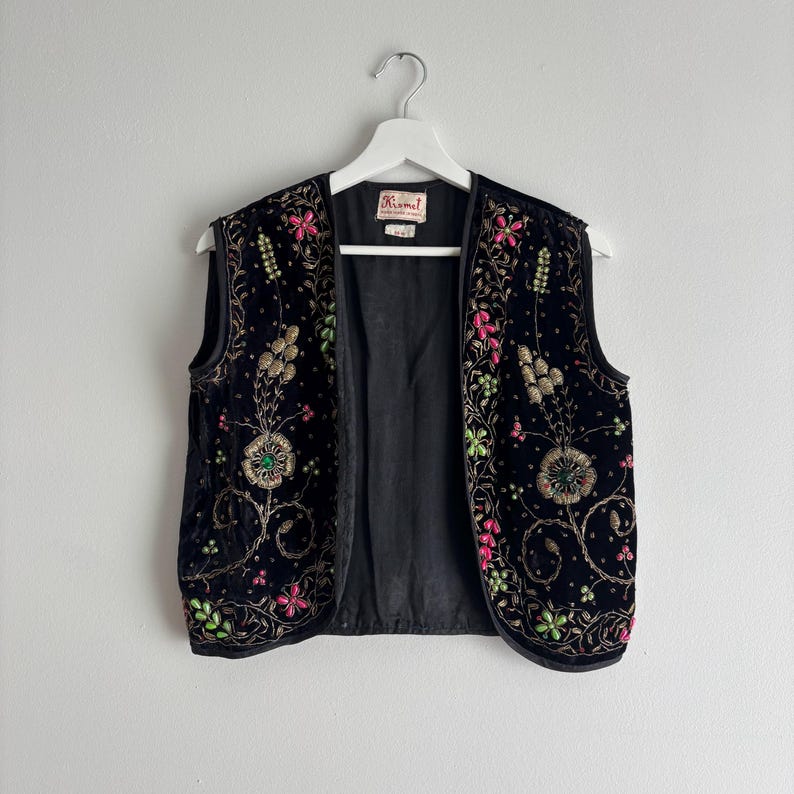 以下が含まれることがあります： 黒いベルベットのベスト、カラフルな花の刺繍入り。 ピンク、緑、金色の刺繍の花と葉が特徴です。 ノースリーブのベストは白いハンガーにかかっています。