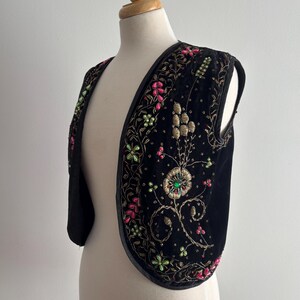 以下が含まれることがあります： ピンク、緑、金色の花の刺繍が施された黒いベルベットのベスト。 ベストは、中央の花とビーズのアクセントを備えた装飾的なデザインが特徴です。 エッジは黒いボーダーでトリミングされており、ヴィンテージのボヘミアンスタイルを作り出しています。