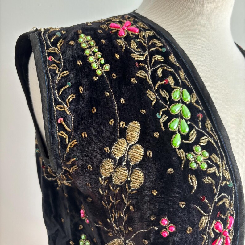 以下が含まれることがあります： 黒いベルベットのベストで、複雑な金の刺繍とカラフルな花のアクセントが特徴です。 ベストはVネックのデザインで、ピンク、緑、金色の装飾が施されています。 刺繍は、ベストの表面にテクスチャード加工された華やかなパターンを作り出しています。