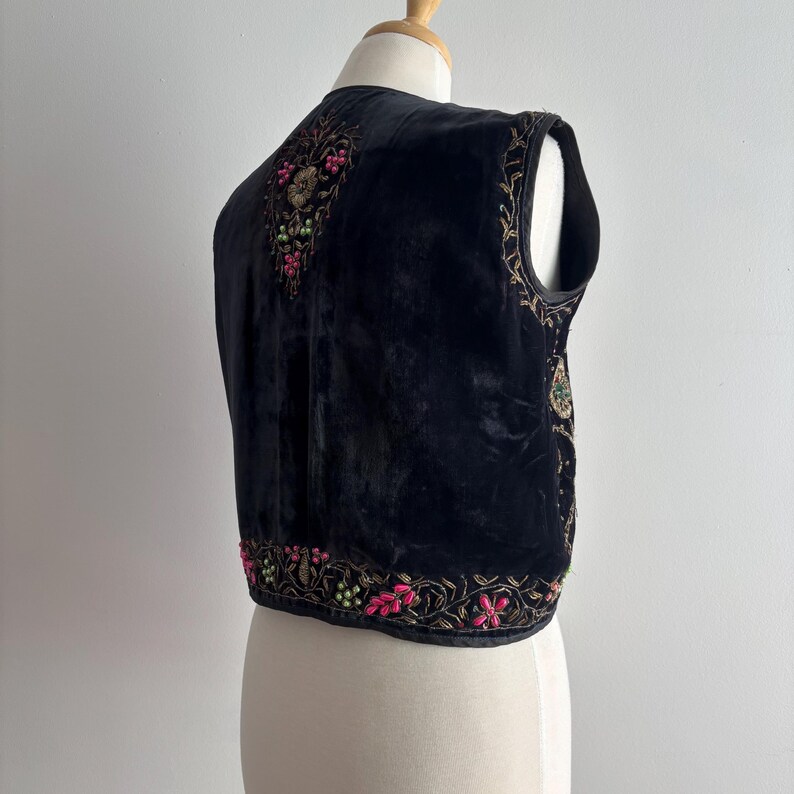 以下が含まれることがあります： ピンク、ゴールド、グリーンで複雑な花の刺繍が施された黒いベルベットのベスト。 ベストは、ヴィンテージまたはボヘミアンスタイルを示す、縁とネックラインに沿ったテーラードフィットと詳細な装飾が特徴です。