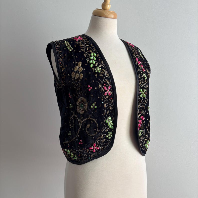 以下が含まれることがあります： 黒いベルベットのベスト。金、ピンク、緑色の花の刺繍が施されています。ノースリーブのベストはVネックのデザインです。刺繍は詳細な花のパターンと小さなビーズのアクセントが特徴で、装飾的な衣服を作り出しています。