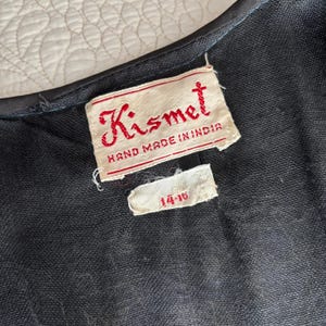 以下が含まれることがあります： 黒い衣服のクローズアップ。赤い文字で「Kismet HAND MADE IN INDIA」と書かれたラベルが付いています。小さなタグにはサイズ範囲「14-16」が表示されています。生地は暗く、質感のある素材のようです。