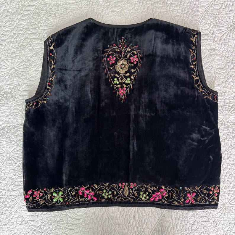 以下が含まれることがあります： ピンク、緑、金色の花の刺繍が施された黒いベルベットのベスト。 ベストは背中にハート型のデザインと縁に花のディテールが特徴です。 ベストはノースリーブで、丸いネックラインです。