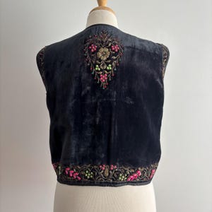以下が含まれることがあります： ピンク、緑、金色の花の刺繍が施された黒いベルベットのベスト。 ベストは背中にハート型のデザインと、縁に花のディテールが施されています。 ベストはマネキンに展示されています。