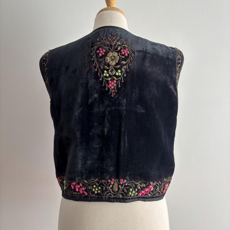以下が含まれることがあります： ピンク、緑、金色の花の刺繍が施された黒いベルベットのベスト。 ベストは背中にハート型のデザインと、縁に花のディテールが施されています。 ベストはマネキンに展示されています。