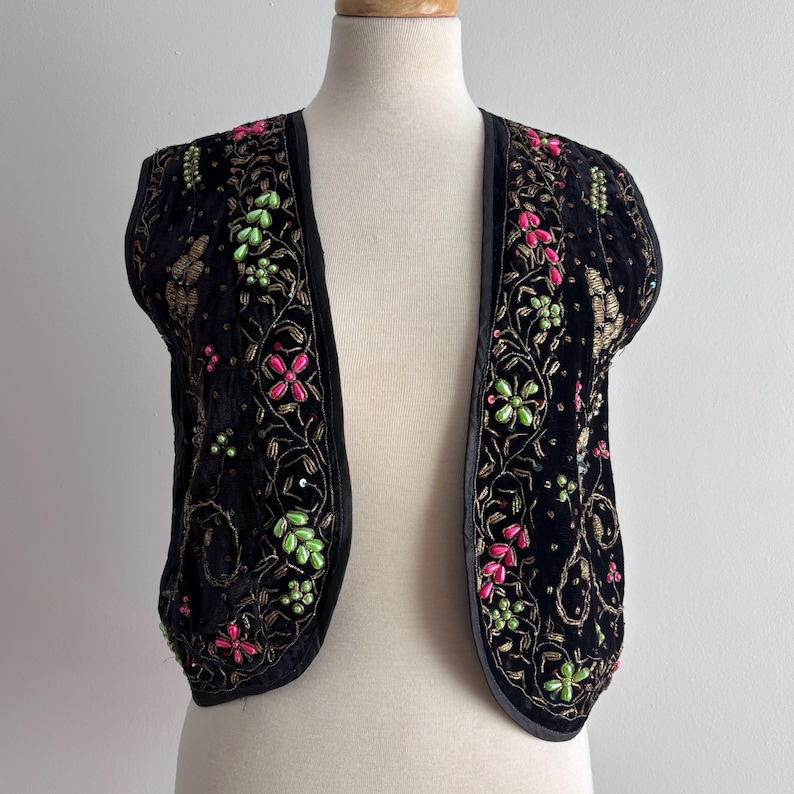 以下が含まれることがあります： ピンク、グリーン、ゴールドの花柄刺繍が施された黒いベルベットのボレロジャケット。 オープンフロントのジャケットはサテンのトリムが特徴で、マネキンに展示されています。 刺繍にはスパンコールと詳細な花柄が含まれています。
