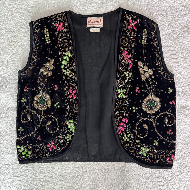 以下が含まれることがあります： ピンク、緑、金色の花の刺繍が施された黒いベルベットのベスト。ノースリーブのベストは丸いネックラインです。刺繍は詳細な花と葉が特徴です。ベストには「Kismet」と書かれたラベルが付いています。