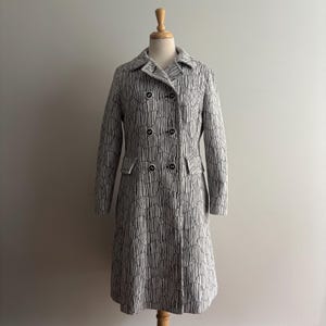 May include: A vintage double-breasted coat in a black and white pattern. The coat features a notched collar, two rows of black buttons, and two front pockets. The coat is displayed on a wooden mannequin.
