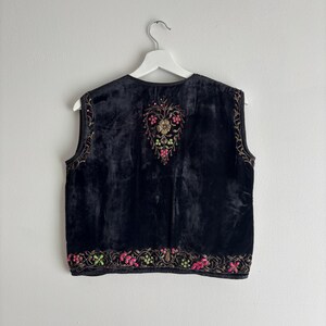 以下が含まれることがあります： ピンク、緑、金色の花の刺繍が施された黒いベルベットのベスト。ノースリーブのベストは丸いネックラインで、白いハンガーに吊るされています。刺繍はベストの前面と背面に集中しています。