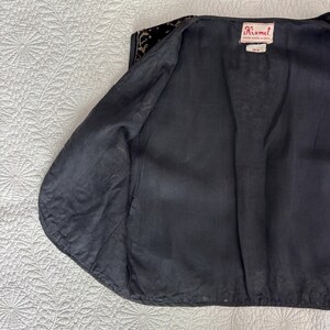 以下が含まれることがあります： 花柄の刺繍と濃い青色の裏地が付いた、開いた黒いベスト。「Kismet hand made jewels」と書かれたラベルが付いています。ベストは開いた状態で表示され、内側の裏地が見えます。