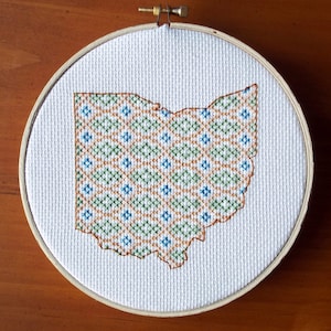 Pode incluir: Um aro de bordado branco com um desenho de ponto cruz do estado de Ohio. O desenho apresenta um padrão geométrico em tons de verde, azul e laranja.