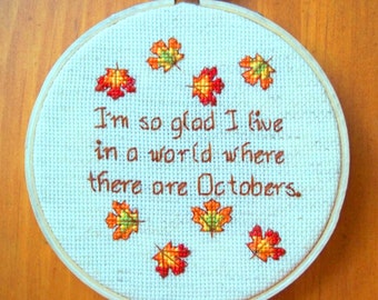 May include: Embroidered hoop art with a blue background and brown text that reads "I'm so glad I live in a world where there are Octobers."  The hoop is decorated with orange, yellow, and green maple leaves.