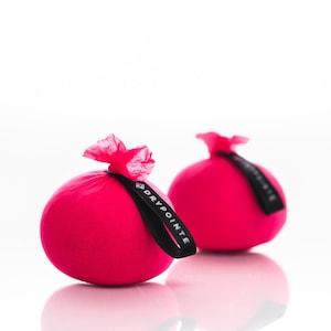 May include: Two pink fabric balls with black ribbon ties and the text "DRYPOINTE" on the ribbon. The balls are on a white surface.