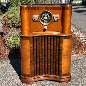 May include: A vintage floor-standing radio cabinet crafted from polished brown wood. The unit features a central circular analog dial with white markings, flanked by horizontal control knobs. The lower section displays vertical wooden slats.