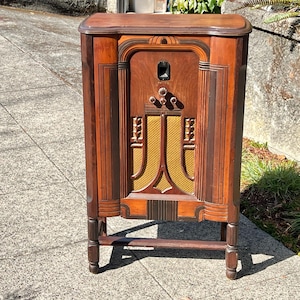 May include: A tall, rectangular wooden radio cabinet featuring a dark brown finish with vertical grooved detailing. The front panel includes a central gold-colored speaker mesh, four circular control knobs, and a small rectangular display window.