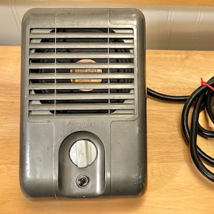 May include: A vintage gray electric heater with a black power cord. The heater has a grill, a dial, and the brand name "Sparkle" on the front. The power cord is coiled and tied with a red twist tie.