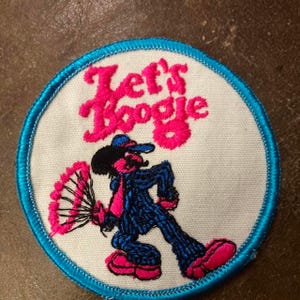 May include: Embroidered patch with the text "Let's Boogie" in pink. The patch features a cartoon figure in a blue suit, pink tie, and pink shoes, holding a pink fan. The patch has a white background with a blue border.