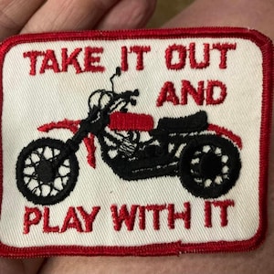 May include: Embroidered patch with a red border and a white background. The patch features a black motorcycle with red accents and the text "TAKE IT OUT AND PLAY WITH IT" in red.