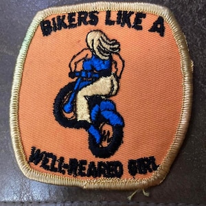 May include: Embroidered patch with the text "BIKERS LIKE A WELL-BEARDED GIRL". The patch features a woman riding a motorcycle, with blue and gold accents on an orange background. The patch has a gold border.