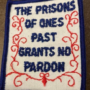 May include: Embroidered patch with the text "THE PRISONS OF ONES PAST GRANTS NO PARDON" in blue thread on a white background. The patch has a blue border and red decorative embroidery.