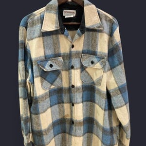May include: A vintage plaid shirt in shades of blue, cream, and gray. The shirt features a classic button-down design with two chest pockets and a black collar. The label reads "McGregor".