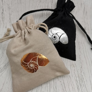 May include: Two drawstring pouches, one beige and one black, are displayed. The beige pouch features a gold nautilus shell design, while the black pouch has a silver nautilus shell. Both pouches are made of fabric and have a drawstring closure.