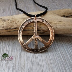 May include: A copper peace sign pendant necklace made from a 1918 British penny. The pendant is on a black cord and is resting on a piece of driftwood.