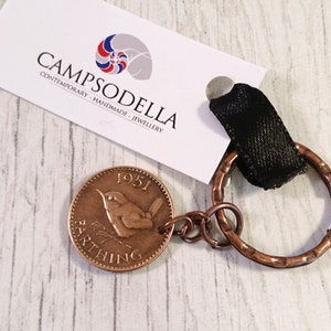 May include: A copper Farthing coin from 1951, featuring a bird, attached to a copper key ring with a black fabric loop. The coin is part of a keychain. The brand name "CAMPSODELLA" is printed on a white tag.