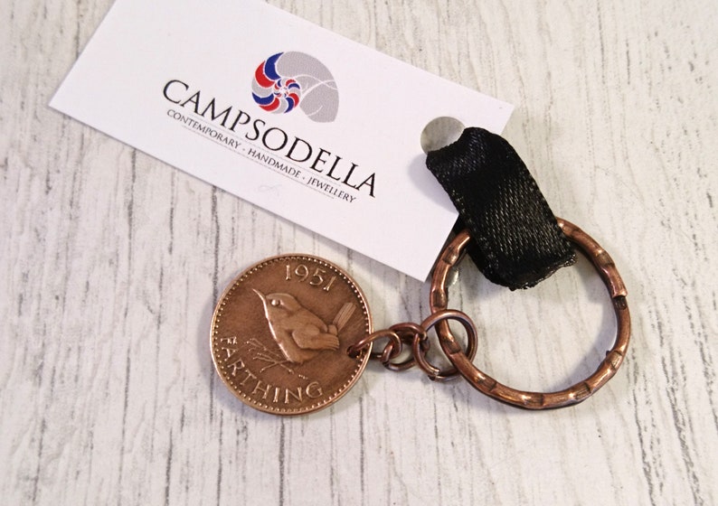 May include: A copper Farthing coin from 1951, featuring a bird, attached to a copper key ring with a black fabric loop. The coin is part of a keychain. The brand name "CAMPSODELLA" is printed on a white tag.