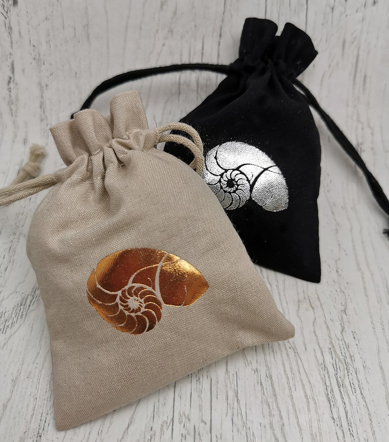 May include: Two small fabric drawstring bags with a nautilus shell design. One bag is beige with a gold shell and the other is black with a silver shell.