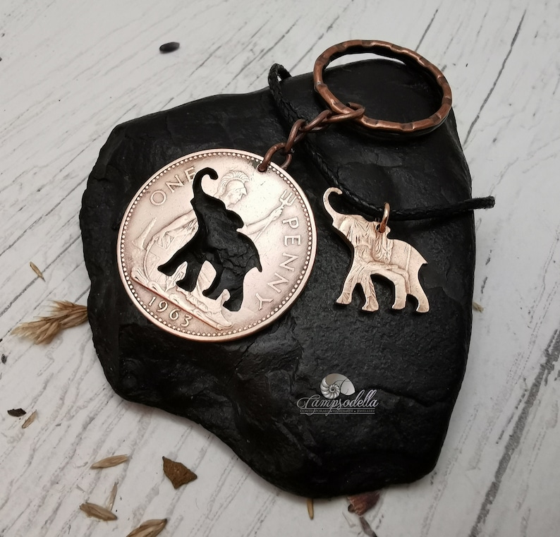 May include: A copper-colored elephant charm hangs from a black cord attached to a key ring. The charm is made from a 1963 British one penny coin with an elephant cut out of the center. The coin is on a black rock.