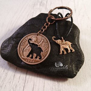 May include: A copper-colored key chain with a 1948 British One Penny coin featuring an elephant silhouette. A smaller elephant charm hangs from a black cord.
