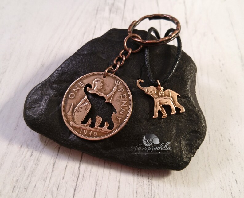 May include: A copper-colored key chain with a 1948 British One Penny coin featuring an elephant silhouette. A smaller elephant charm hangs from a black cord.