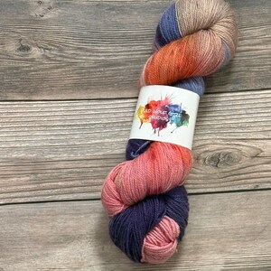 May include: A skein of colorful yarn with shades of blue, orange, pink, and tan. The yarn is wrapped with a white label that reads "BAD WOLF GIRL STUDIOS" in a colorful, artistic font. The yarn is displayed on a wooden surface.