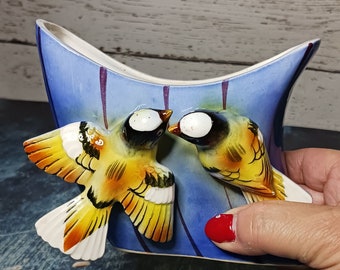 May include: A blue and white ceramic vase with two yellow birds perched on the rim. The birds have black eyes and white accents.